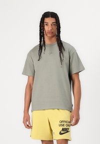 Olijf groene katoenen T-shirt met korte mouwen en een ronde halslijn, met een klein geborduurd Nike-logo op de borst. Gele shorts met zwarte tekst.