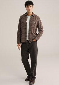 Ruda languota marškinėliai su dviem krūtinės kišenėmis, baltais mygtukais, dėvimi ant šviesiai pilko marškinėlio, derinami su tamsiomis kelnėmis ir rudais batais.