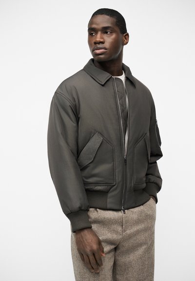 Mannequin masculin portant une veste bomber gris foncé avec poches avant et poignets côtelés, associée à un pantalon beige texturé, debout sur un fond blanc.
