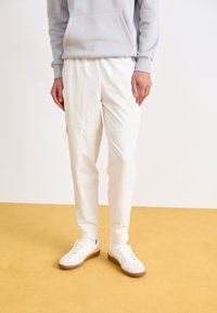 Vita joggerbyxor med en slät yta, med en dragkedjeficka på låret. Matchas med vita sneakers och en ljusgrå hoodie.