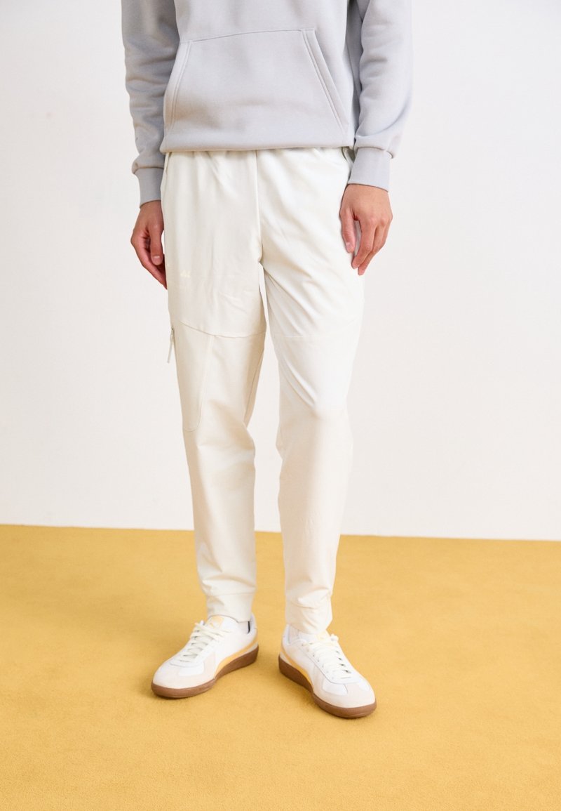 Witte joggingbroeken met een gladde textuur, voorzien van een ritszak op de bovenbeen. Gepaard met witte sneakers en een lichtgrijze hoodie.