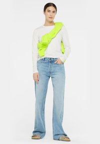 Langärmliges weißes Hemd, hellblaue weite Jeans und ein leuchtend neongrüner Strickpullover, der über die Schulter gelegt ist. Legere Schuhe sind ebenfalls enthalten.