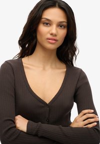 Brun ribbet sweater med en dyb V-hals, lange ærmer og delikat bølget kant. Har tre sorte knapper foran. Glat tekstur.