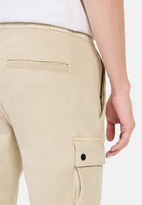 Beige Cargo-Hosen mit elastischem Bund, Seitentaschen und einer hinteren Taschen mit Knopf. Der Stoff wirkt glatt und strapazierfähig.