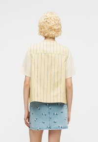 Personne aux cheveux blonds courts et bouclés portant un haut à manches courtes rayé jaune et blanc et une jupe en jean bleue avec de petites fleurs brodées.