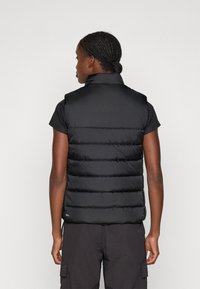 Vest negru pufos fără mâneci, cu guler înalt, model cusut orizontal și textură netedă, asociat cu un top închis la culoare cu mâneci scurte.