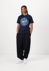 Marineblå kortermet t-skjorte med en fargerik grafisk logo i midten, kombinert med løse svarte bukser og hvite sneakers.