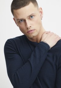 Muška mornarska košulja dugih rukava, s uskim krojem. Na njegovom zapešću nalazi se srebrna narukvica, koja se kontrastira s teksturom košulje.