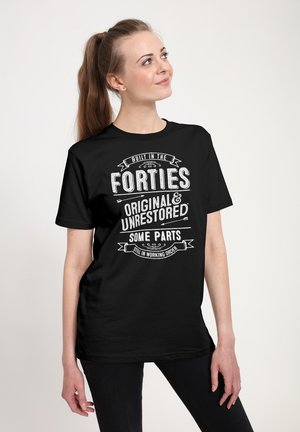 Kvinde med langt hestehale iført en sort T-shirt med hvid tekst om at være "Bygget i fyrreerne," stående foran en ensfarvet baggrund.