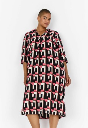 Žena v volném šatě délky po kolena s růžovým, černým a bílým geometrickým vzorem, stojící před jednobarevným bílým pozadím.