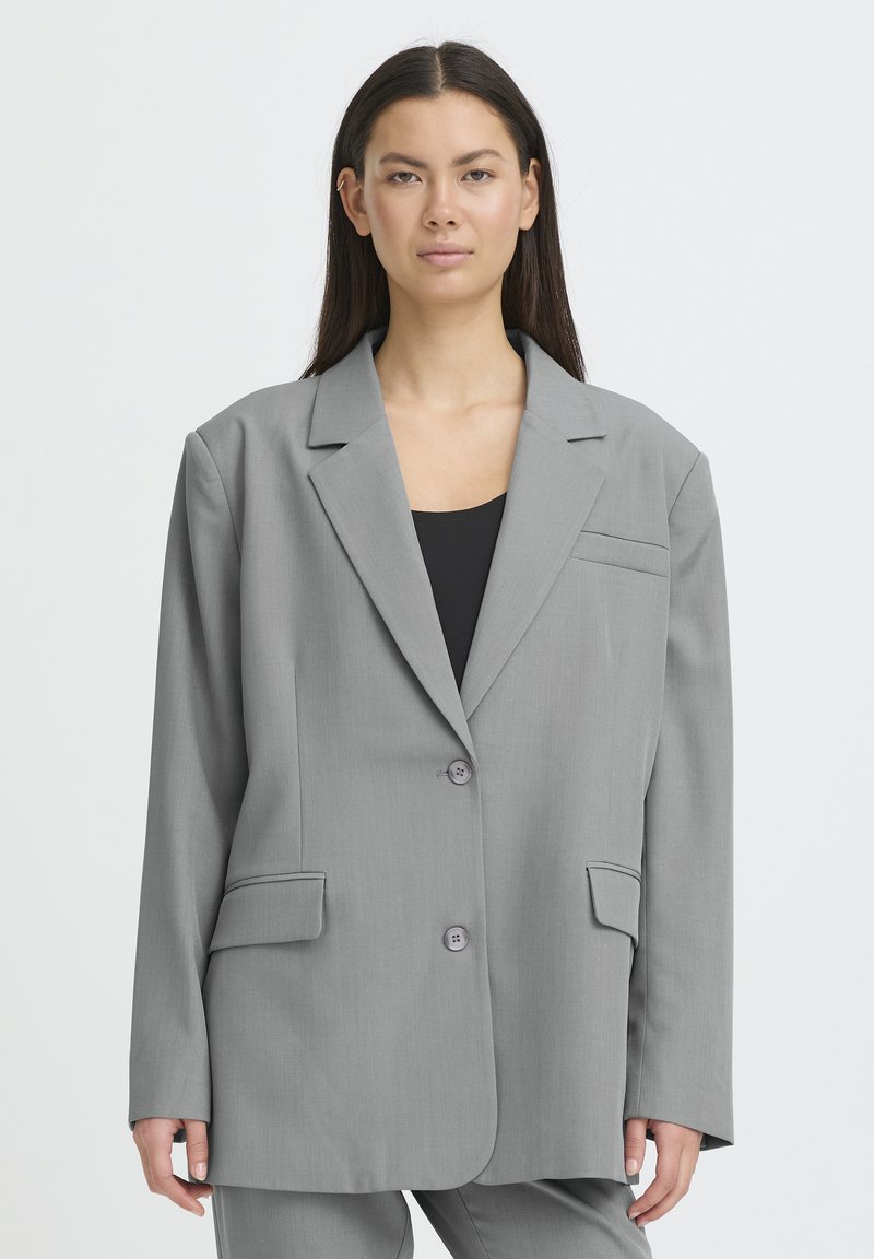 Femme aux longs cheveux foncés portant un blazer oversize gris clair et un pantalon assorti sur un haut noir, debout devant un fond clair uni.