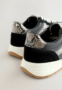 Melni sporta apavi ar teksturētu čūsku raksta papēdi un zamšādas akcentiem. Baltā zole ar gumijas apakšu, kas izceļ pelēkus un melnus akcentus.