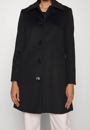 Žena v dlouhém černém kabátu se čtyřmi knoflíky, doplněném světlými kalhotami, stojící před jednobarevným pozadím.