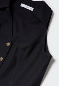 Chemise noire sans manches avec col, présentant une texture lisse et trois fermetures à boutons à l'avant. Étiquette de la marque visible au niveau de l'encolure.