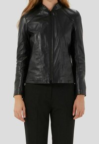 Veste en cuir noir au design ajusté, avec un col haut et une fermeture éclair à l'avant. Présente des lignes épurées et une texture lisse. Associée à un pantalon noir.