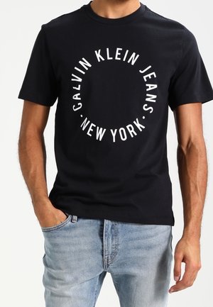 Mann trägt schwarzes T-Shirt mit weißem, rundem „Calvin Klein Jeans New York“-Schriftzug, kombiniert mit hellblauen Jeans.