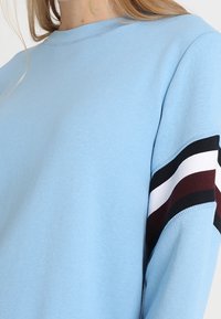 Felpa azzurra con righe orizzontali nere, bianche e rosso scuro sulla parte superiore della manica, indossata da una persona con capelli biondi.