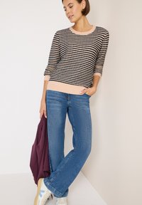 Gebreid vest in zwart met roze stippen, met geribbelde zoom, gecombineerd met lichtblauwe jeans en witte sneakers. Houdt een bordeauxrode jas vast.