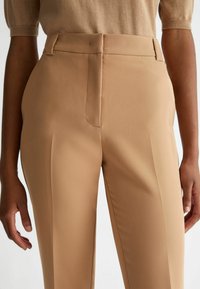 Beige op maat gemaakte broeken van gladde stof, met een rechte snit, subtiele plooien aan de voorkant en zijzakken.