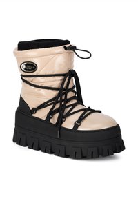 Beżowy but z lakierowanej skóry z czarną gumową podeszwą, grubym bieżnikiem, wyściełanym kołnierzem i czarnymi sznurowadłami. Posiada naszywkę z logo po boku.