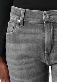 Gros plan sur la ceinture et la poche d’un jean gris porté avec un haut noir côtelé, montrant le bouton, la boucle de ceinture et les détails cousus sur le tissu.