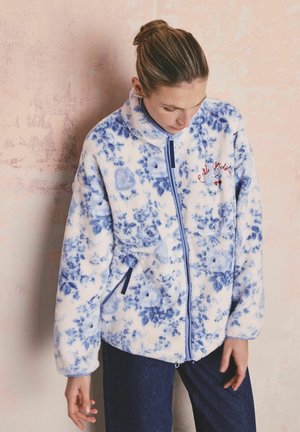 Žena v bielej fleecovej bunde s modrými kvetinovými vzormi stojí pred štruktúrovanou svetloružovou stenou a pozerá sa dole.