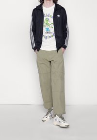 Svart träningsjacka med vita ränder, grafisk vit T-shirt, olivgröna cargobyxor och mönstrade sneakers med en ljus färgpalett.