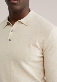 Mann trägt beige langärmliges Poloshirt mit zwei Knöpfen, dargestellt von den Schultern bis zur Brustmitte vor einem einfarbigen Hintergrund.