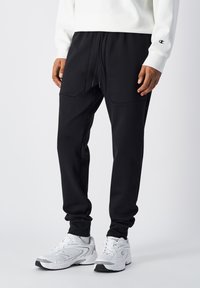 Zwarte joggingsbroek met een relaxte pasvorm, voorzien van twee voorzakken en een koordsluiting in de taille, gecombineerd met witte sport schoenen.
