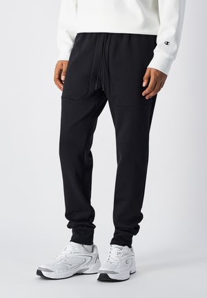 Black jogger pants with a relaxed fit, featuring two front pockets and a drawstring waistband, paired with white athletic shoes.