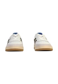 Sneakers bianchi con strisce verdi, punta rotonda, chiusura con lacci, texture morbida e suola in gomma beige, caratterizzati da una linguetta blu e dettagli perforati.