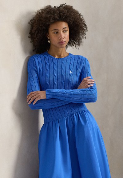 Μπλε πουλόβερ με πλέξη καλτσοδέτας και μακριά μανίκια, συνδυασμένο με μια αέρινη μπλε φούστα. Σημαντικές λεπτομέρειες περιλαμβάνουν εφαρμοστή μέση και υφασμάτινη υφή.