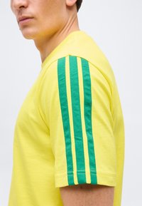 Άνδρας που φοράει κίτρινη κοντομάνικη μπλούζα με τρεις κάθετες πράσινες ρίγες στο μανίκι, εμφανιζόμενος από το πλάι πάνω σε απλό φόντο.