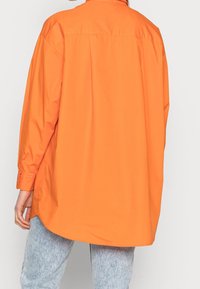 Persona che indossa una maglietta a maniche lunghe oversize di un vivace arancione e jeans leggeri in lavaggio acido blu, mostrata di spalle.