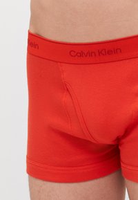 Crvene pamučne bokserice s širokim elastičnim pojasom na kojem je otisnut logo "Calvin Klein". Prilagođen dizajn s prednjim otvorom.