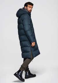 Lange, puffy donkergroene parka met een capuchon, voorzien van horizontale naden, een ritsafsluiting en zijknopen. Gepaarde met donkergroene broek en zwarte laarzen.