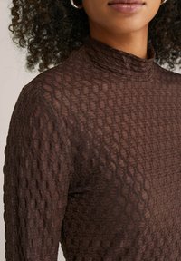 Ženska z kodrastimi lasmi, ki nosi srebrne obroče za ušesa in teksturirano rjavo dolgotrajno majico z visokim ovratnikom in dolgimi rokavi.