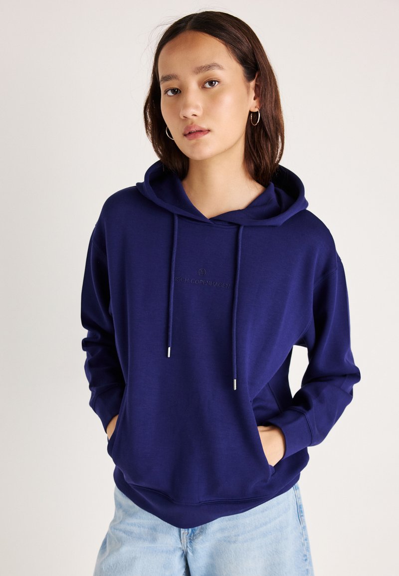 Námornícky modrá mikina s kapucňou a šnúrkami, s prednou kapsou a jemným výšivkovým logom. Mäkkučký materiál a voľný strih.