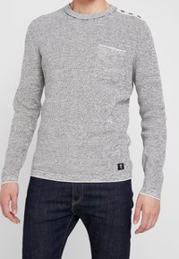 Grau-weißer Texturoberteil mit Rundhalsausschnitt, langen Ärmeln, einer Brusttasche und Knopfdetails an der Schulter. Getragen mit dunklen Jeans.