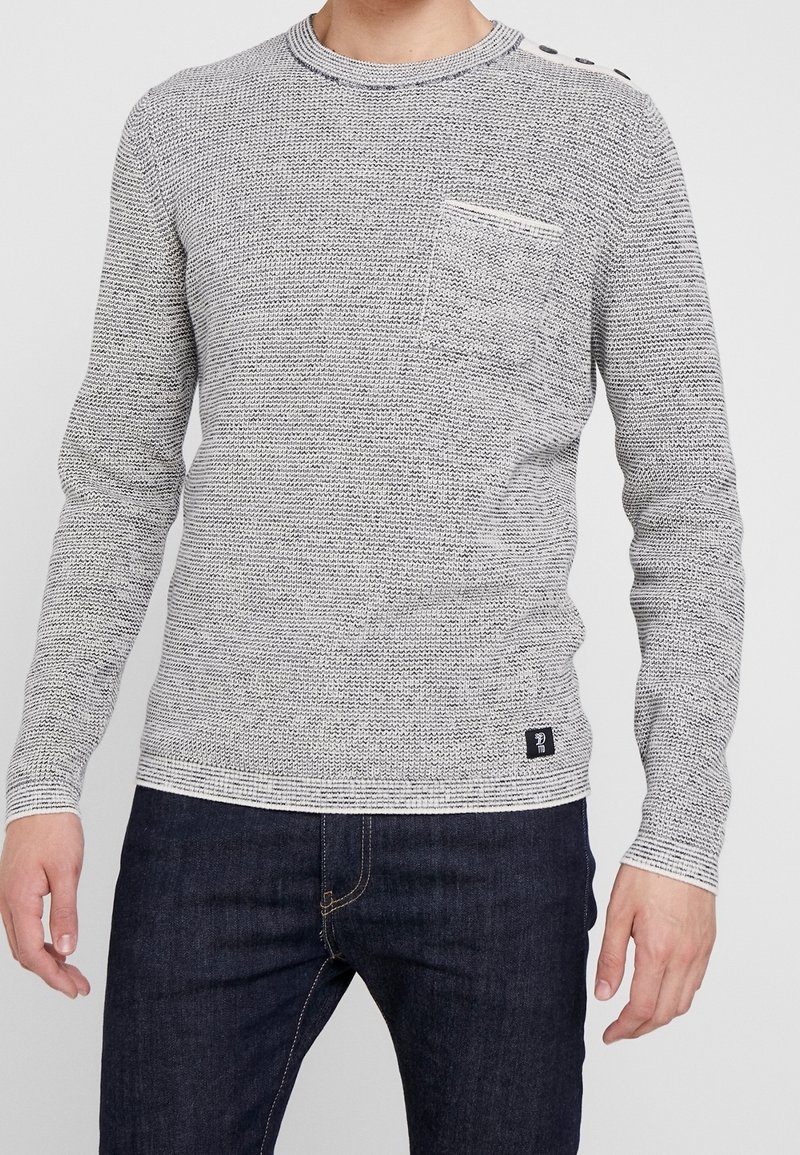 Grau-weißer Texturoberteil mit Rundhalsausschnitt, langen Ärmeln, einer Brusttasche und Knopfdetails an der Schulter. Getragen mit dunklen Jeans.