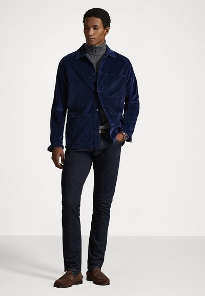 Ναυτικό καμπαρντίνα από κοτλέ με κουμπιά στο μπροστά μέρος, δύο τσέπες και υφή, φορεμένη πάνω από ένα γκρι πουλόβερ και σκούρους τζιν. Καφέ λοφάκια.