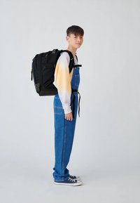 Black backpack worn on the back, featuring multiple zippers and a sturdy design. Light orange to white gradient sweatshirt with blue denim overalls.