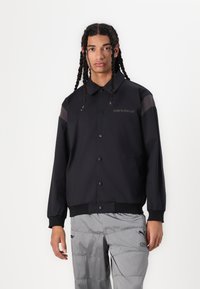 Chaqueta negra de manga larga con cuello, que presenta paneles transparentes en contraste y el logo de "New Balance" en el pecho. Textura de tejido suave.
