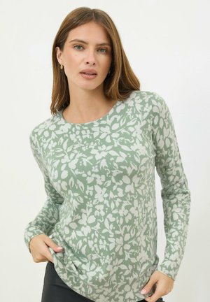 Frau mit glattem, hellbraunem Haar, die einen hellgrünen Pullover mit weißem Blumenmuster trägt, Hände in Hüfthöhe ruhend, neutraler Hintergrund.