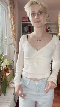 Jovem com cabelo loiro curto, suéter branco, jeans claros e tatuagem no peito com a palavra "ART" está dentro de casa ao lado de uma janela e de uma estante.