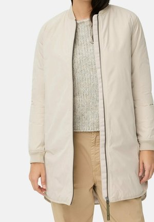 Personne portant une longue veste zippée beige clair sur un pull gris en maille et un pantalon beige, debout devant un fond uni.