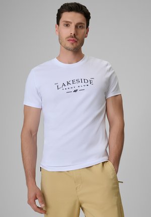 Mężczyzna noszący białą koszulkę z napisem "Lakeside Yacht Club" i beżowe spodnie, stojący na jednolitym szarym tle.
