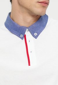 Άνδρας που φοράει λευκό πόλο με μπλε γιακά με κουμπιά και κόκκινο περιθώριο κατά μήκος της σειράς κουμπιών, κοιτάζει προς τα δεξιά.