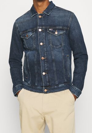 Homme portant une veste en jean bleu foncé avec des boutons à l’avant et un pantalon beige, mains dans les poches, sur un fond blanc uni.