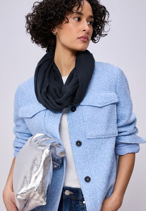 Žena s kudrnatými vlasy má na sobě světle modrý texturovaný kabát, černý šátek a drží velkou kovovou stříbrnou kabelku.
